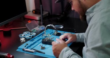 Masasında tişörtle oturan bir adamın FPV insansız hava aracının video verici çipini lehimlediği orta ölçekli bir fotoğraf. Genç bir mühendis bir elektronik panoyu lehimliyor. Akşamları adam hobisiyle meşgul oluyor..