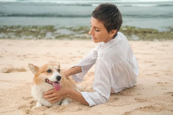 A middle-aged woman in a white shirt with a short haircut strokes her Welsh Corgi on the beach. The dog lies on the sand with its tongue hanging out. Traveling with a pet. Banner pet store.