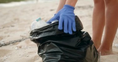 Gönüllüler sahildeki çöpleri temizliyor. Plastik dolu bir çöp torbasının yakın çekimi. Eldiven takan bir kadın plastik şişe takıyor. Çevreye önem vermek.