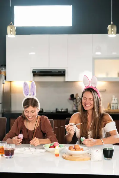 Mom and daughter wearing hair holders with bunny ears sit at the table and decorate Easter eggs. They are smiling and laughing. Celebrating Easter with family. The main spring Christian holiday.