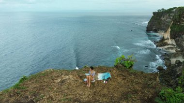 Okyanus kıyısındaki bir uçurumun kenarında oturan ve tuvale resim çizen bir kadın ressamın havadan görünüşü. Bir deniz manzarası çiziyor. Elinde bir palet tutuyor ve mavi boyaları fırçayla karıştırıyor..
