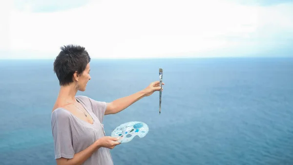 Portrait of an artist against the backdrop of the ocean, who extends her hand with a painting brush in front of her and continues to mix paints. The artist paints a landscape on the ocean from life.