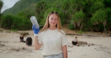 Genç bir kadın plajı plastik çöplerden temizliyor, elinde plastik bir şişe var, onu çantaya koyuyor ve parmağıyla sahile çöp atmanın yasak olduğunu gösteriyor.