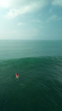 Bir sörfçünün sörf tahtasının üzerinde yüzdüğü okyanus manzarasının dikey videosu. Kırmızı mayo giymiş genç ve seksi bir kadın sörf tahtasında yatarken sırada kürek çekiyor. Yüksek dalgalarda sörf yapmak.