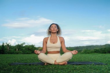 Kıvırcık saçlı genç bir Latin kadın yoga minderinde nilüfer pozisyonunda oturur ve şafak vakti yeşil bir parkın arka planında meditasyon yapar. Nefes ve zihin konsantrasyonu. Farkındalık pratiği.