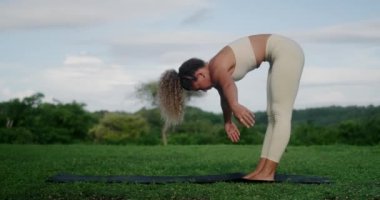 Genç ve çekici bir Latin atletik kadın yeşil bir ormanın arka planında yoga yapıyor ve derin derin nefes alıyor. Yoga eğitmeni şafakta parkta güneş selamı veriyor.