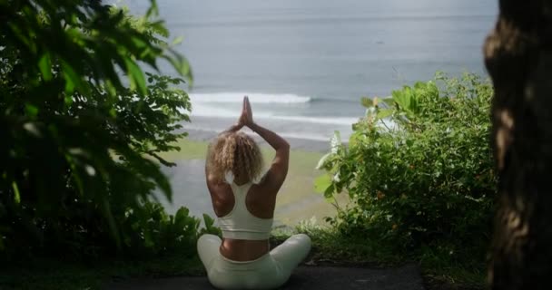 蓮の位置の崖の端に座っているカーリーヘアの若い女性の後ろからの眺めは 海を見て瞑想します 心の平和 静けさ そして静けさ — ストック動画