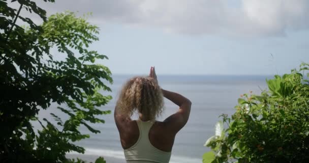 一个卷发的年轻女子坐在悬崖边上的荷花式的位置上 从她的后面看去 她望着大海 沉思着 心灵的平静 沉默和自然的平静 — 图库视频影像
