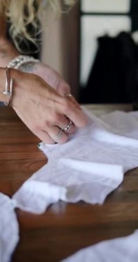 Beyaz pamuktan bir tişört diken bir kadın terzinin elleri dikey videoda, kadın iğnelerle kumaş parçalarını birleştiriyor. Tasarımcı çevre dostu kumaşlardan elbiseler üretir..