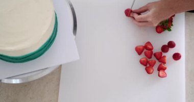Pasta dükkanındaki bir masanın çalışma yüzeyi. Beyaz bir kesim tahtasındaki pasta süslemesi için taze böğürtlen kesen genç bir pasta şefinin ellerine yakın çekim.