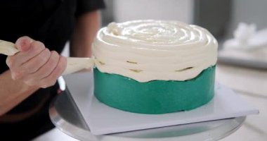 Genç bir pasta şefinin, pikaptaki bir pastanın yüzeyine beyaz krema sürerken yakın plan çekimleri. Bir şekerleme dükkanında üretim sürecinde çalışma. Pasta dekorasyonu.
