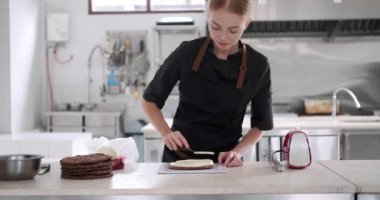 Orta boy, genç, kızıl saçlı bir pasta şefinin pasta poşeti kullanarak çikolatalı pastaya tereyağı sürerken görüntüsü. Profesyonel mutfaktaki bir pastanede pasta hazırlama süreci..