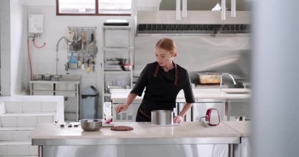 赤毛の若い女性のペストリーシェフが働くペストリーショップでプロのキッチンの長距離ショット 彼女は金型でケーキを準備し 層の1つにバタークリームを広げる — ストック動画