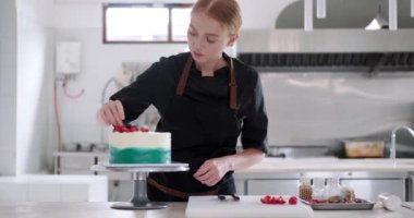 Bir şekerci dükkanındaki parlak bir prodüksiyonun orta ölçekli görüntüsü. Genç kızıl saçlı çekici kadın pasta şefi pastayı taze böğürtlenlerle süslemeye odaklanıyor. Profesyonel çalışma süreci.