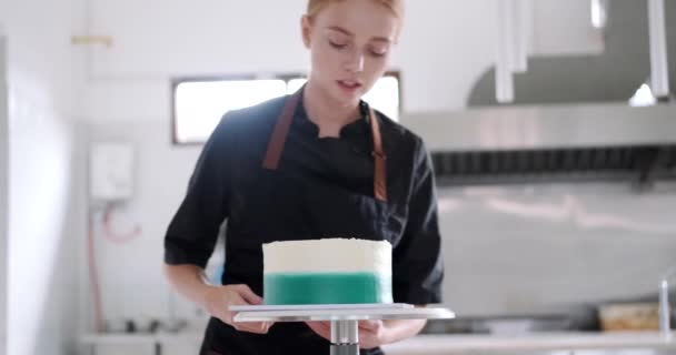 若い魅力的な赤毛の女性のペストリーシェフは ペストリーショップの産業台所のテーブルからケーキを取り 冷蔵庫に持って行きます 甘いデザートを準備するプロセス — ストック動画