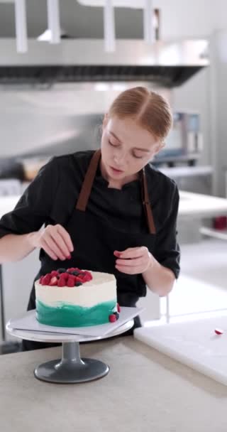 菓子店での明るい生産の垂直ビデオ 若い赤毛の魅力的な女性のペストリーシェフは 新鮮なベリーでケーキを飾ることに集中しています プロフェッショナルな作業プロセス — ストック動画