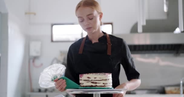レストランのプロのキッチンで赤毛の女性のペストリーシェフは ペストリーバッグからターコイズクリームでケーキを飾ります 菓子店でデザートを準備するプロセス — ストック動画