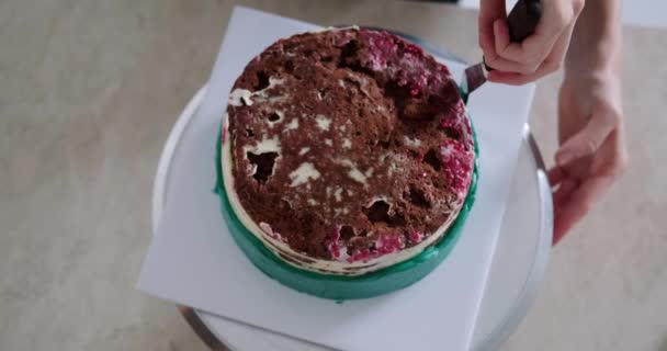 回転スタンドに立っているラズベリー層のチョコレートケーキのトップビュー 若い女性のペストリーシェフはそれの上にターコイズクリームを広げます 菓子店で働くプロセス — ストック動画
