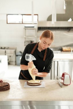 Genç kızıl saçlı bir pasta şefinin portresi pasta poşeti kullanarak çikolatalı pastaya tereyağı sürüyor. Profesyonel mutfaktaki bir pastanede pasta hazırlama süreci..