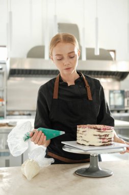 Bir restorandaki profesyonel bir mutfakta genç kızıl saçlı bir pasta şefi pasta çantasından çıkan turkuaz kremayla bir pasta süslüyor. Şekerleme dükkanında tatlı hazırlama süreci..