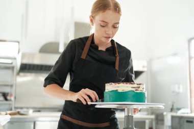 Bir restorandaki profesyonel bir mutfakta genç kızıl saçlı bir pasta şefi pasta çantasından çıkan turkuaz kremayla bir pasta süslüyor. Şekerleme dükkanında tatlı hazırlama süreci..