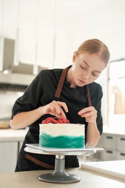 Bir şekerci dükkanındaki parlak bir prodüksiyonun dikey fotoğrafı. Genç kızıl saçlı çekici kadın pasta şefi pastayı taze böğürtlenlerle süslemeye odaklanıyor. Profesyonel çalışma süreci.