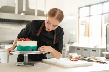 Bir şekerci dükkanındaki parlak bir prodüksiyonun orta ölçekli görüntüsü. Genç kızıl saçlı çekici kadın pasta şefi pastayı taze böğürtlenlerle süslemeye odaklanıyor. Profesyonel çalışma süreci.