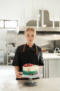 Çekici kızıl saçlı genç bir pasta şefinin portresi. Yapım atölyesinin arka planına karşı önlük giymiş. Masanın üzerinde taze böğürtlenlerle süslenmiş bir pasta var..