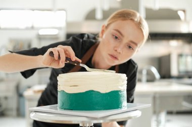 Genç, kızıl saçlı bir pasta şefinin portresi. Pastaya konsantrasyonuyla bakıyor ve kremayı yüzeyine düz bir tabaka halinde dağıtıyor. Bir üretim mutfağında hazırlanma süreci