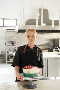 Çekici kızıl saçlı genç bir pasta şefinin portresi. Yapım atölyesinin arka planına karşı önlük giymiş. Masanın üzerinde taze böğürtlenlerle süslenmiş bir pasta var..