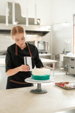 Restoran mutfağındaki genç, kızıl saçlı bir pasta şefi turkuaz krema dağıtıyor. Dönen tezgahta spatula kullanıyor. Şekerleme dükkanında tatlı hazırlama süreci..