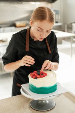 Bir şekerci dükkanındaki parlak bir prodüksiyonun dikey fotoğrafı. Genç kızıl saçlı çekici kadın pasta şefi pastayı taze böğürtlenlerle süslemeye odaklanıyor. Profesyonel çalışma süreci.