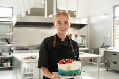 Çekici kızıl saçlı genç bir pasta şefinin portresi. Yapım atölyesinin arka planına karşı önlük giymiş. Masanın üzerinde taze böğürtlenlerle süslenmiş bir pasta var..