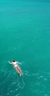 Beyaz bikinili seksi bir sporcu kadının dikey hava videosu. Mükemmel sörf yerine ulaşmak için turkuaz suda sörf tahtasını yüzdürüyor..