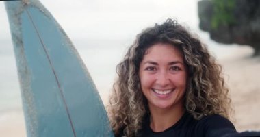 Kıvırcık saçlı ve çilli genç bir Latin sörfçü kameraya selfie çekiyor, elinde sörf tahtası tutuyor ve okyanusun arka planına gülümsüyor. Sevimli bir sörfçünün portresi.