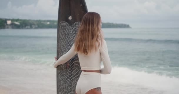 白い水着の若い金髪の女性がビーチに立って 海で波をキャッチしているサーファーの距離を調べ 彼女は彼女の手でサーフボードを取り 水に入ります — ストック動画