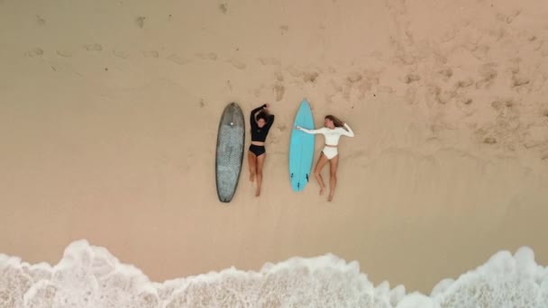 两个穿着比基尼的年轻女冲浪者躺在海滩上 坐在冲浪板旁边的沙滩上 自由快乐的冲浪人们在靠近大海的沙滩上放松 — 图库视频影像