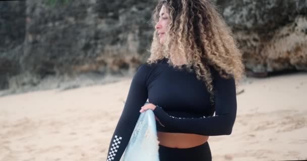 黒いサーフ水着のカーリーヘアを持つ若いセクシーなラティナの女性は ビーチに立って 彼女の手でサーフボードを保持しています 彼女は遠くを見つめ 微笑んでいる サーフィンは生き方である 極端なスポーツの女性 — ストック動画