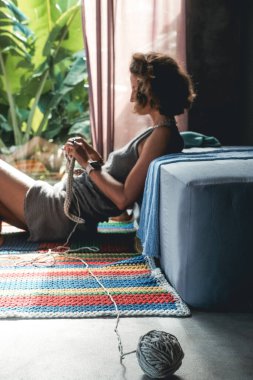 Ön planda, arka planda yünlü bir iplik topu var. Kısa saçlı genç bir kadın yerde oturuyor ve yanında bir sepet aletle battaniye örüyor..