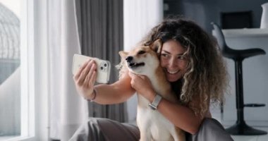 Kıvırcık saçlı genç bir kadın Shiba Inu köpeğiyle selfie çekiyor. Köpeğe sarılıyor ve kameraya bakarken gülümsüyor. Sosyal medyada arkadaşça ve arkadaşça bir sevgiyle çekilmiş bir fotoğraf..