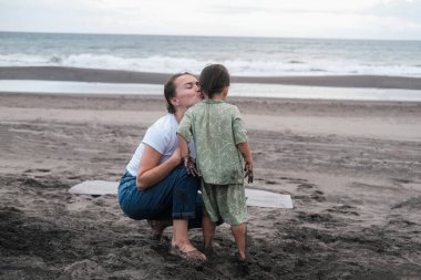 Kumsalda bir anne yanağından öpüyor. Yanında duran küçük oğlunu. Sıcak bir tropik ülkede tatilde olan bir çocukla yaz tatili. Aile içinde sevgi ve şefkat. Anneler Günü