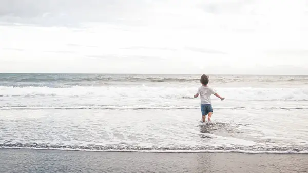 A happy toddler boy is running joyfully into the ocean water on the beach, under the blue sky and fluffy clouds. The liquid and fluid waves splash around him as the wind gently blows. Mothers day.