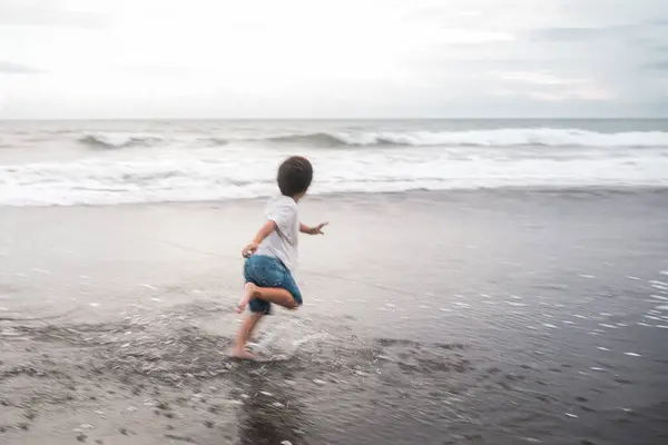 A happy toddler boy is running joyfully into the ocean water on the beach, under the blue sky and fluffy clouds. The liquid and fluid waves splash around him as the wind gently blows. Mothers day.