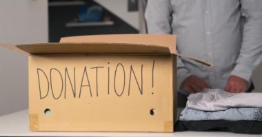 Üzerinde yazılı bağış olan karton bir kutunun durduğu masanın yakın çekimi. Bir adam bir kutuya çeşitli günlük giysiler koyar. Hayır işi için bir şeyler topluyorum. Düşük gelirli ailelere yardım.