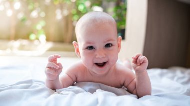 Arka bahçede, yeşil yaprakların olduğu bir arka planda, 4 aylık bir bebek karnının üzerinde battaniyeye uzanmış, kameraya bakıyor ve duygusal olarak gülüyor. Mutlu yeni doğanlar anneleriyle oynuyor..