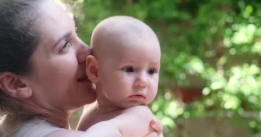 Anne, bebeği kollarında tutuyor ve yanağından öpüyor. Anneler Günü. Arka bahçede annesiyle birlikte yeni doğmuş küçük bir kız. Arka planda yeşil ağaçlar, çocuklarla yaz yürüyüşleri.