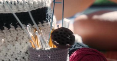 Yanında çok renkli yün ipliklerden oluşan bir sepetin yakın çekimi iğneler ve kancalarla dolu bir bardakta duruyor. Bir kadın eli örgü iğnelerini seçer ve onları camdan çıkarır..