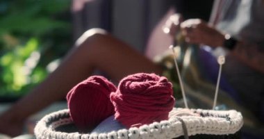 Genç bir kadın yatak odasında yerde oturuyor ve yün ipliklerden bir battaniye örüyor, ön planda yün topları ve iğneli bir bardak ile örülmüş bir sepet var. Meditasyon hobisi örgü örmek.
