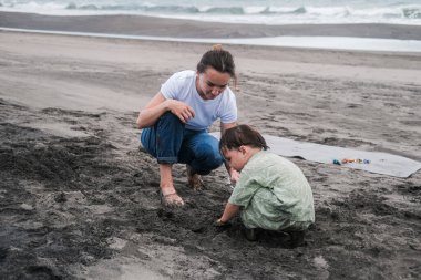 Anne ve oğlu günlük giysiler içinde okyanusun yakınındaki kumsalda otururken oynuyorlar. Çocuğunla yaz tatilinin tadını çıkarıyorsun. Küçük bir çocuk kum kazıyor. Anneler Günü.
