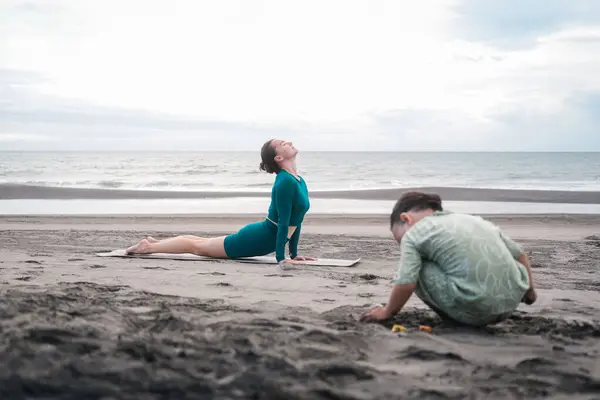 Młoda Kobieta Dresie Uprawia Jogę Plaży Pobliżu Oceanu Jej Synek Obrazek Stockowy